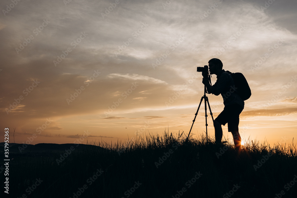 Silhouette of competent photographer with tripod and digital camera taking sunset photos while standing on high hill. Mature man in summer outfit enjoying favorite hobby on fresh air.