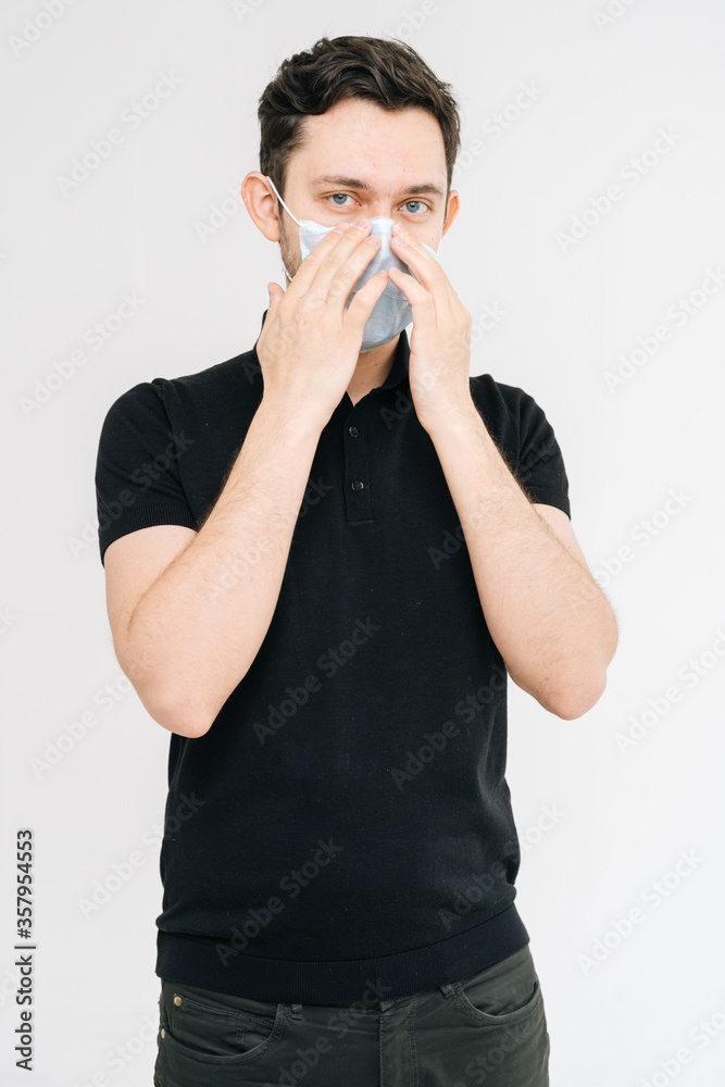 Portrait of serious young man in medical mask wearing casual black shirt, looking at camera, on white background. Prevention of virus infection. Concept of Coronavirus COVID-19 Pandemic. Portrait of serious young man in medical mask wearing casual black shirt, looking at camera, on white background. Prevention of virus infection. Concept of Coronavirus COVID-19 Pandemic.