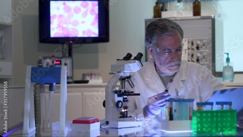 A cancer research scientist in a laboratory looking at a blood sample and taking notes with display of cancer cells on the monitor in the background.