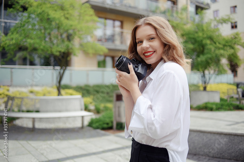 Photographer girl walks around the city with a reflex camera. A young blonde learns to take pictures. The model is looking at the camera and holding a camera in her hands. Photo Freelance Smile