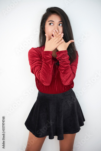 Vertical medium long studio portrait of attractive young Asian woman wearing dark-red top and black skirt covering her mouth in shock, white background