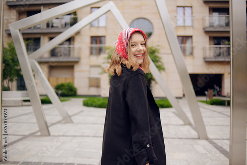 A girl in a business suit with a red scarf on her head against the background of a building, turns to the camera and laughs. Fashion photo of a blonde model poses, in a suit and with a red scarf.