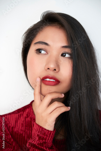 Vertical close-up studio portrait shot of beautiful young Asian woman with dark-red lips thinking of something, white background