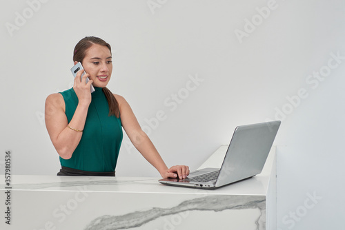 Recepcionista realizando llamada telefónica por celular mientras revisa su laptop, en un cubículo con acabado en mármol y muros de color blanco.