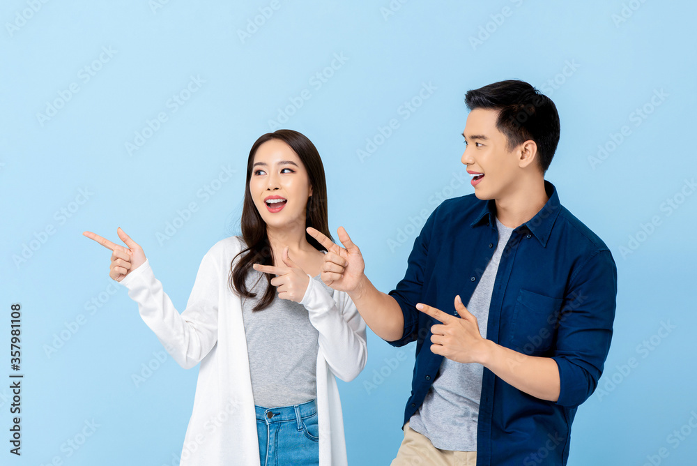 Smiling excited Asian couple tourists pointing hand to empty space on isolated light blue background Smiling excited Asian couple tourists pointing hand to empty space on isolated light blue background
