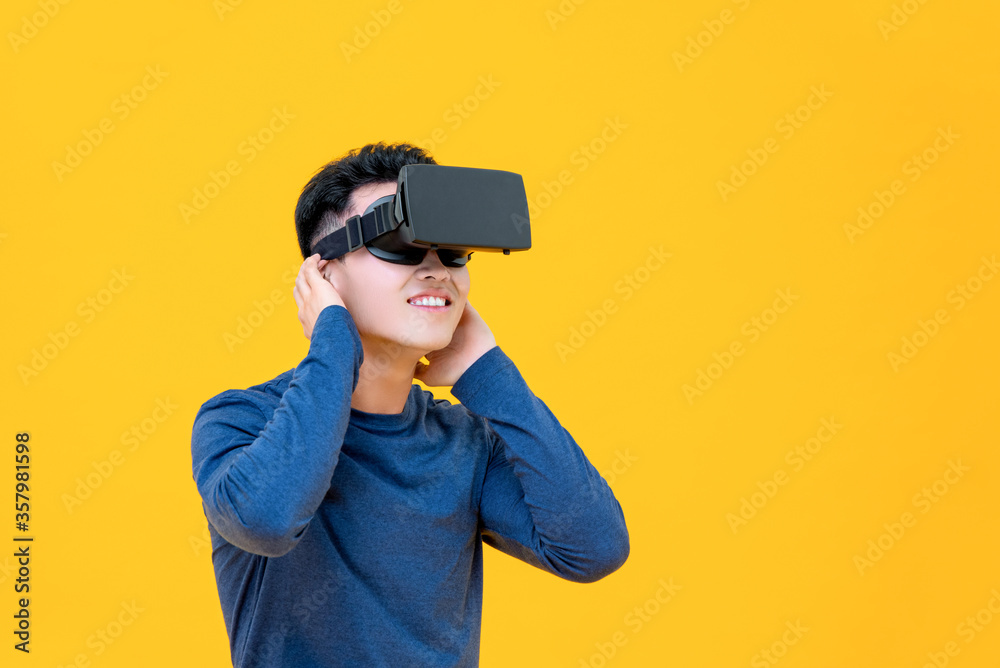 Excited Asian man enjoying watching 3D simulation video from virtual reality or VR glasses on yellow background Excited Asian man enjoying watching 3D simulation video from virtual reality or VR glasses on yellow background