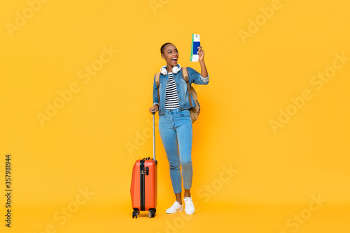 Young excited solo African American woman tourist with baggage passport and boarding pass ready to fly isolated on yellow studio background