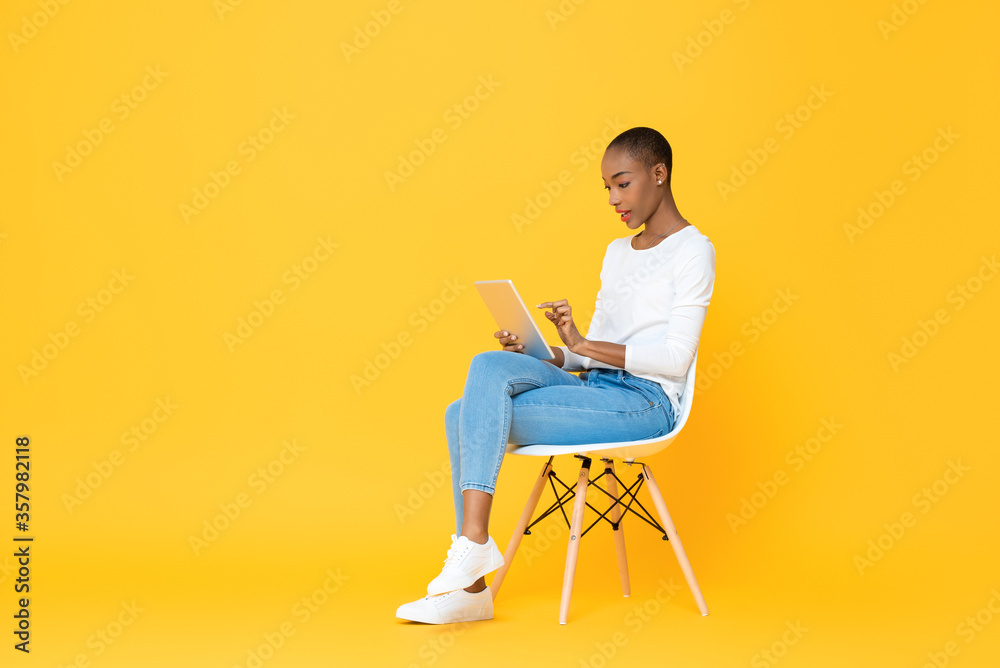 Young African American woman sitting and using tablet computer on isolated yellow background