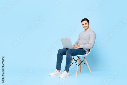 Smiling handsome man sitting on a chair working on laptop computer isolated on light blue background
