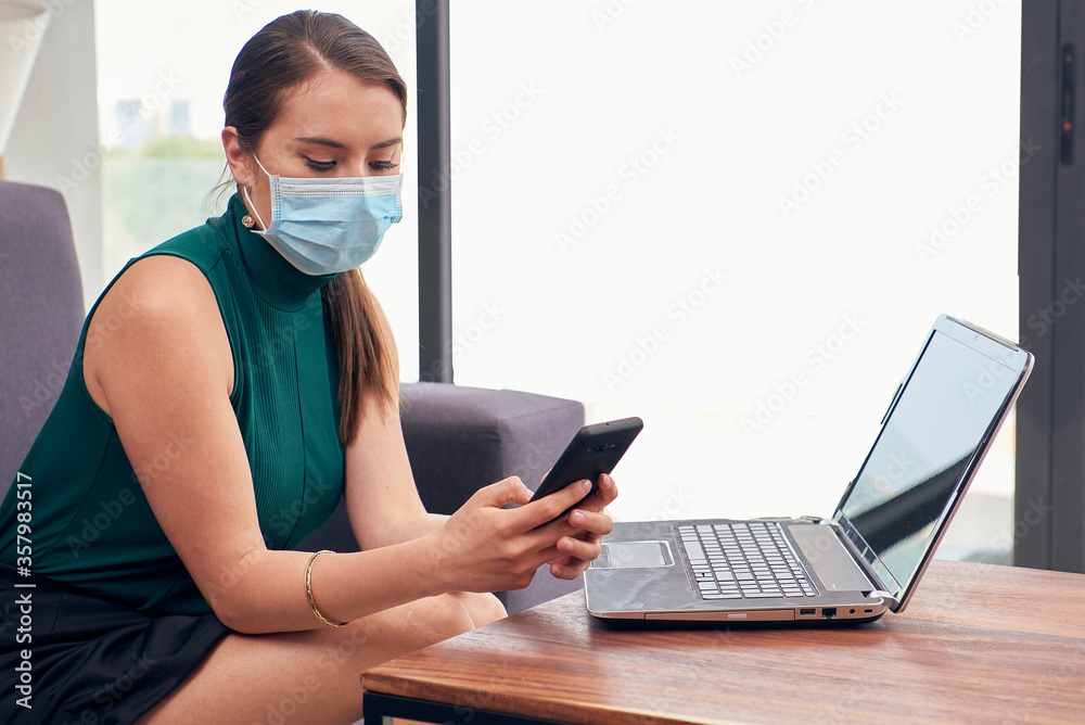Mujer con cubre bocas contestando mensajes en el celular desde casa debido a la nueva normalidad por la pandemia de covid-19 Mujer con cubre bocas contestando mensajes en el celular desde casa debido a la nueva normalidad por la pandemia de covid-19