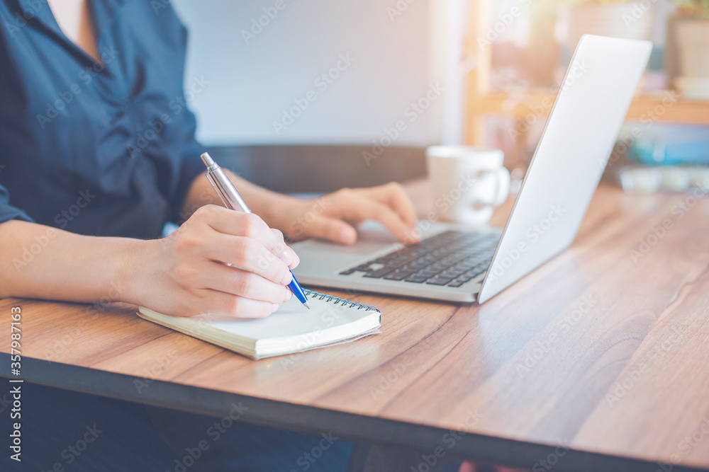 Business woman is writing on a notebook with a pen and using a laptop to work in the office.