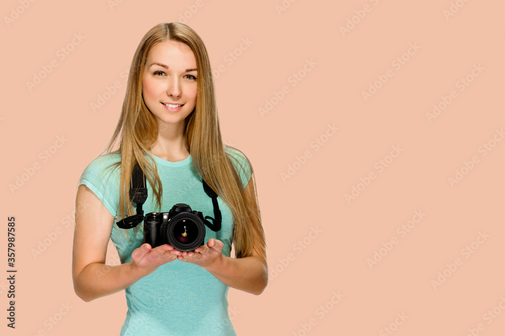 Smiling blonde holding professional photo camera on salmon red background