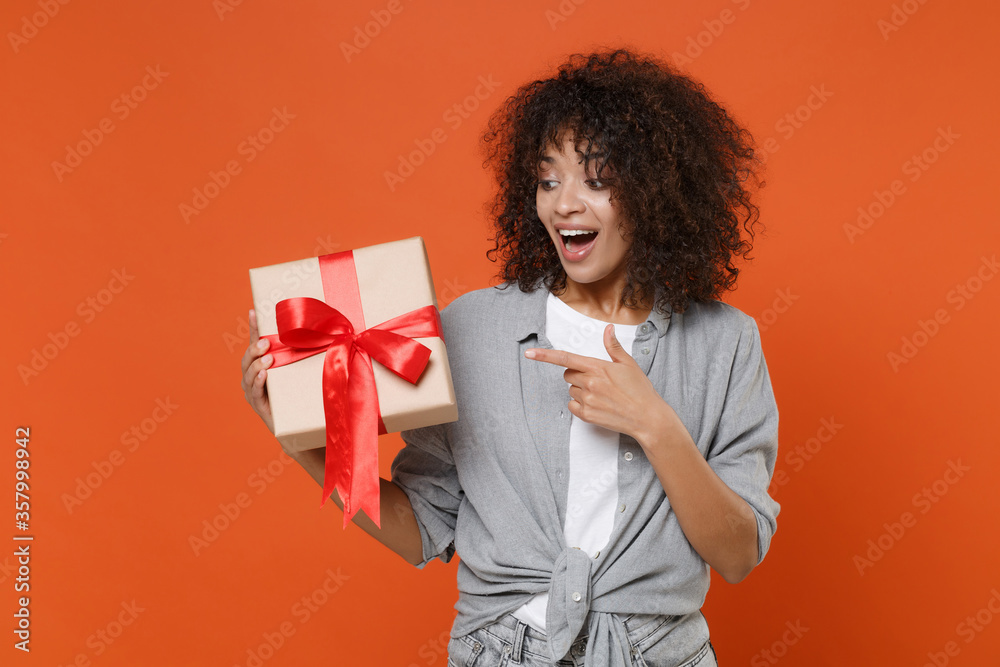 Amazed young african american woman in gray casual clothes isolated on orange background. Valentine's Day Women's Day birthday, holiday concept. Point index finger on present box with gift ribbon bow. Amazed young african american woman in gray casual clothes isolated on orange background. Valentine's Day Women's Day birthday, holiday concept. Point index finger on present box with gift ribbon bow.
