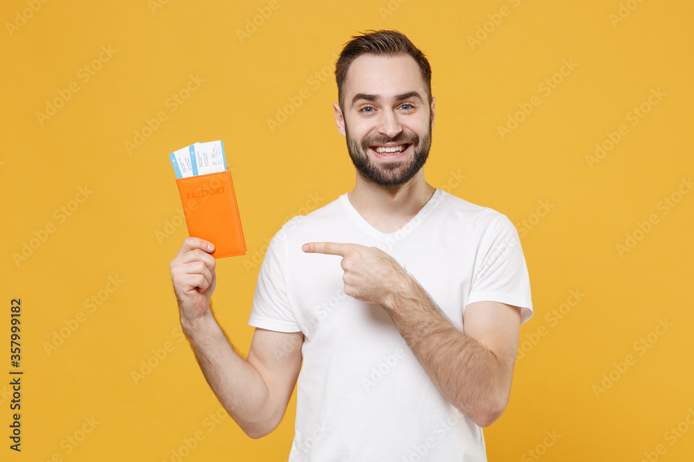 Smiling young man guy in white t-shirt isolated on yellow background. Passenger traveling abroad to travel on weekends getaway. Air flight journey concept. Pointing index finger on passport tickets. Smiling young man guy in white t-shirt isolated on yellow background. Passenger traveling abroad to travel on weekends getaway. Air flight journey concept. Pointing index finger on passport tickets.