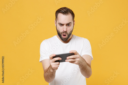 Shocked young bearded man guy in white casual t-shirt posing isolated on yellow wall background studio portrait. People emotions lifestyle concept. Mock up copy space. Play game with mobile phone.