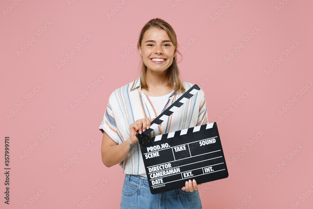 Smiling young blonde woman girl in casual striped shirt posing isolated on pink background studio portrait. People lifestyle concept. Mock up copy space. Hold classic black film making clapperboard.