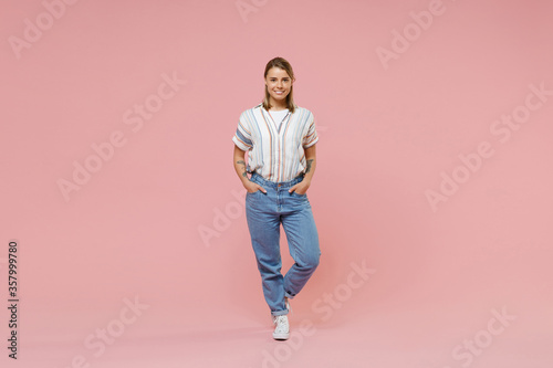 Smiling young blonde woman girl in casual striped shirt posing isolated on pastel pink wall background studio portrait. People emotions lifestyle concept. Mock up copy space. Holding hands in pockets.