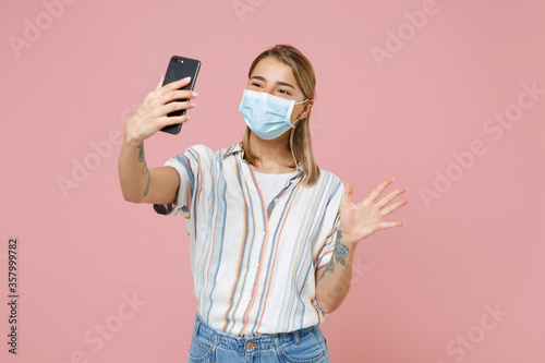 Young woman girl in casual striped shirt sterile face mask isolated on pink wall background. Epidemic pandemic coronavirus 2019-ncov sars covid-19 flu virus concept. Doing selfie shot on mobile phone.