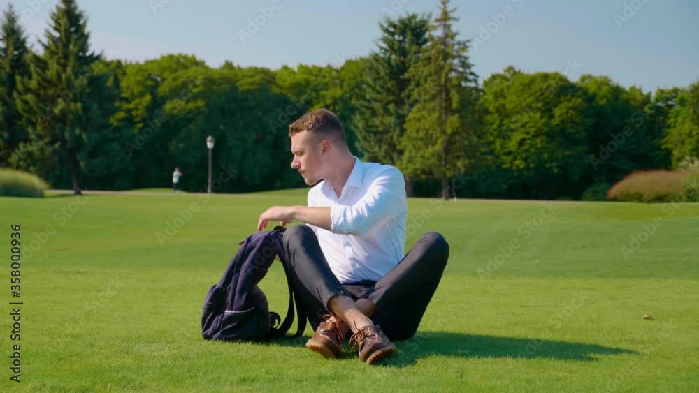 Businessman sits in a park on green grass, takes out vegetarian lunch from backpack and eats. Guy holds an ecological paper package with food and wooden sticks in his hands Businessman sits in a park on green grass, takes out vegetarian lunch from backpack and eats. Guy holds an ecological paper package with food and wooden sticks in his hands