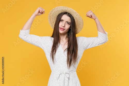 Strong young brunette woman girl in white dress hat posing isolated on yellow wall background studio portrait. People sincere emotions lifestyle concept. Mock up copy space. Showing biceps, muscles.