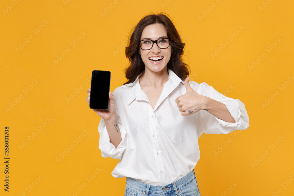 Funny young brunette business woman in white shirt glasses isolated on yellow background studio. Achievement career wealth business concept. Hold mobile phone with blank empty screen showing thumb up. Funny young brunette business woman in white shirt glasses isolated on yellow background studio. Achievement career wealth business concept. Hold mobile phone with blank empty screen showing thumb up.