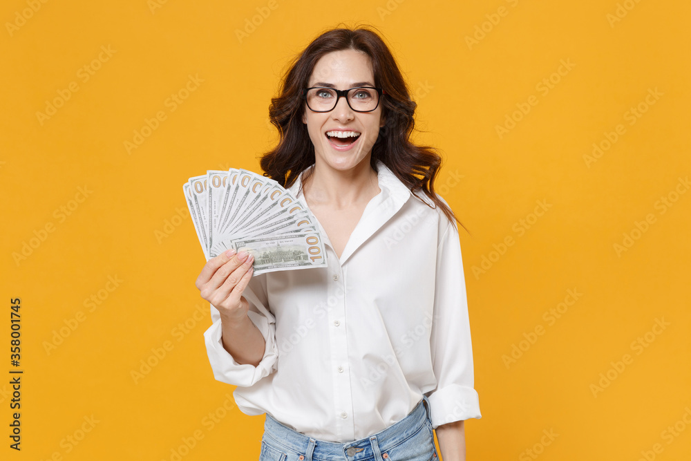 Cheerful young brunette business woman in white shirt glasses isolated on yellow background. Achievement career wealth business concept. Mock up copy space. Hold fan of cash money in dollar banknotes. Cheerful young brunette business woman in white shirt glasses isolated on yellow background. Achievement career wealth business concept. Mock up copy space. Hold fan of cash money in dollar banknotes.