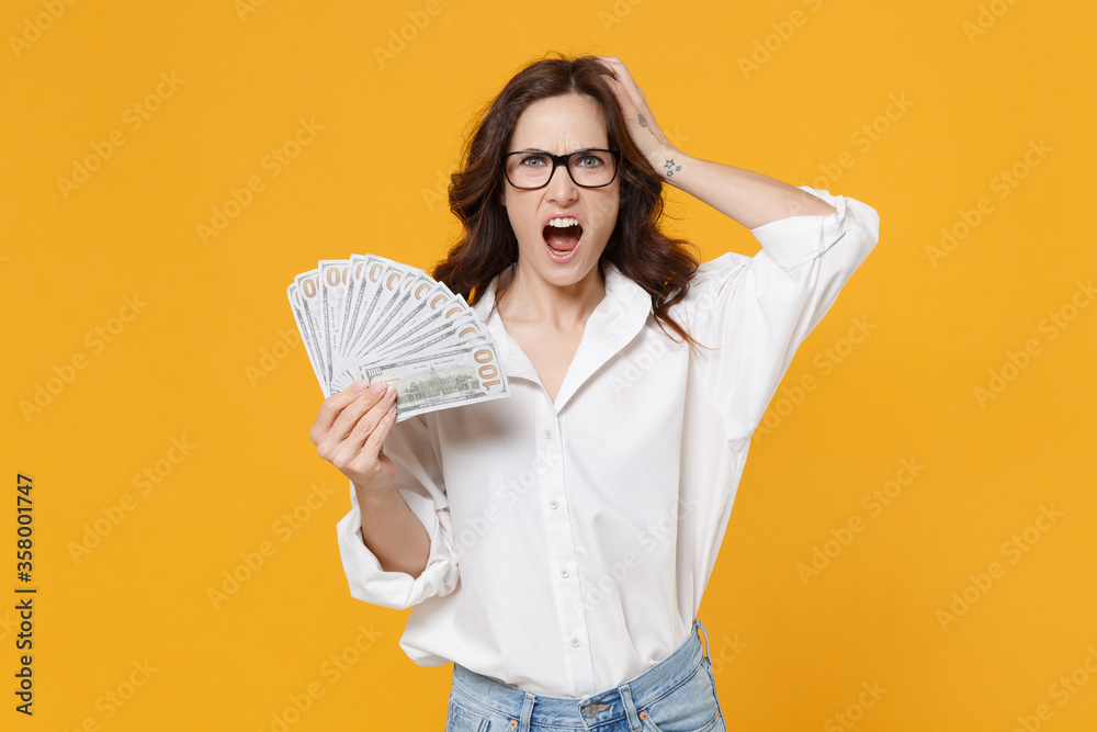 Irritated young brunette business woman in white shirt glasses isolated on yellow background. Achievement career wealth business concept. Hold fan of cash money in dollar banknotes, put hand on head. Irritated young brunette business woman in white shirt glasses isolated on yellow background. Achievement career wealth business concept. Hold fan of cash money in dollar banknotes, put hand on head.
