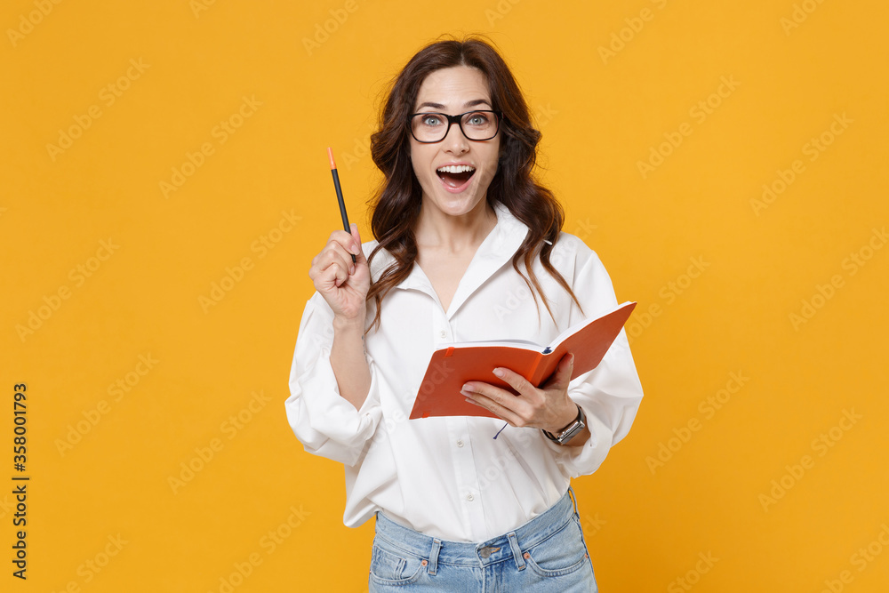 Excited young business woman in white shirt glasses isolated on yellow wall background. Achievement career wealth business concept. Mock up copy space. Hold notebook, point pen up with great new idea. Excited young business woman in white shirt glasses isolated on yellow wall background. Achievement career wealth business concept. Mock up copy space. Hold notebook, point pen up with great new idea.