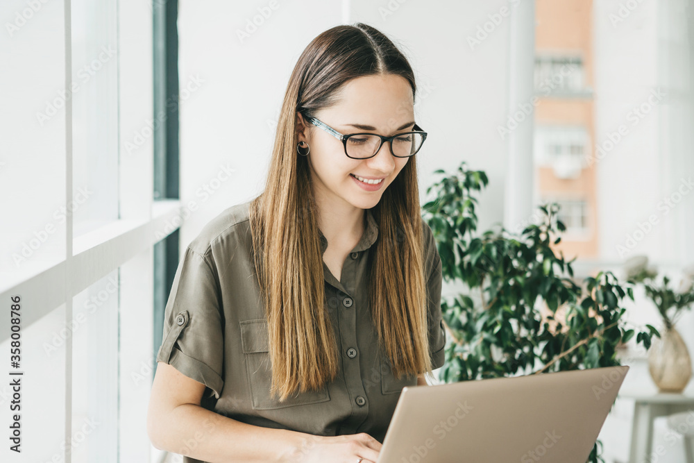 Beautiful young adult positive girl uses a laptop at home by the window or is she in open space. Beautiful young adult positive girl uses a laptop at home by the window or is she in open space.