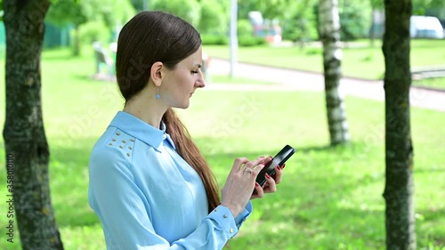 Video Caucasian brunette girl with long hair talking on the phone in the park. A woman dials numbers on the phone, then talks and smiles. Model posing against a background of trees in sunny weather. photo