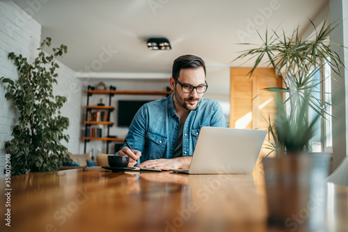 Foto Handsome man taking notes and looking at laptop, at home office, portrait