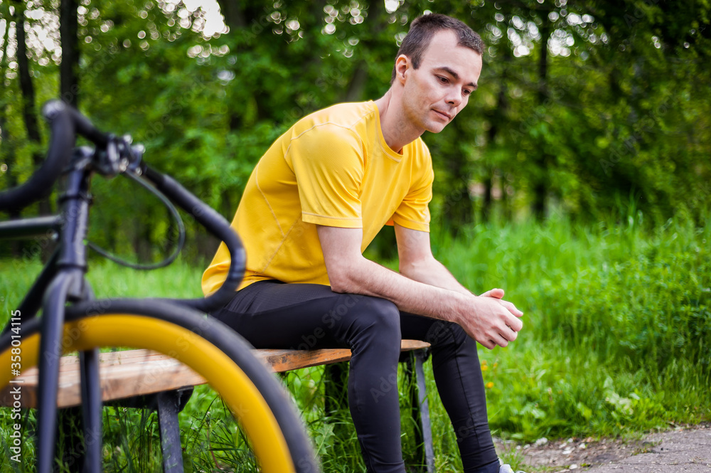 A young man sitting on a bench in a public Park communicates via mobile communication. In the background with his bike. Among trees and vegetation.