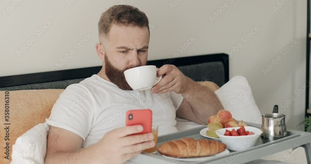 Handsome bearded man using his smartphone while drinking coffee in morning . Guy in 30s looking at phone screen while having breakfast and sitting with tray full of food on bed. Handsome bearded man using his smartphone while drinking coffee in morning . Guy in 30s looking at phone screen while having breakfast and sitting with tray full of food on bed.