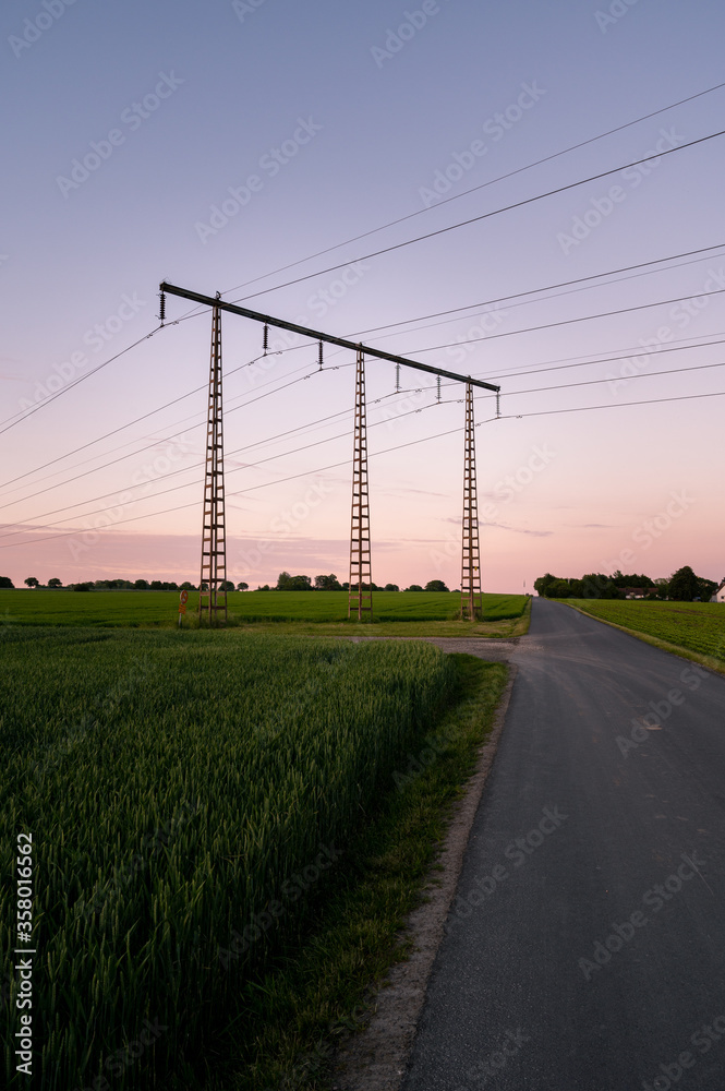 Electricity pylons close to a rural road with green crops growing during summer sunset in Skåne (Scania), Sweden