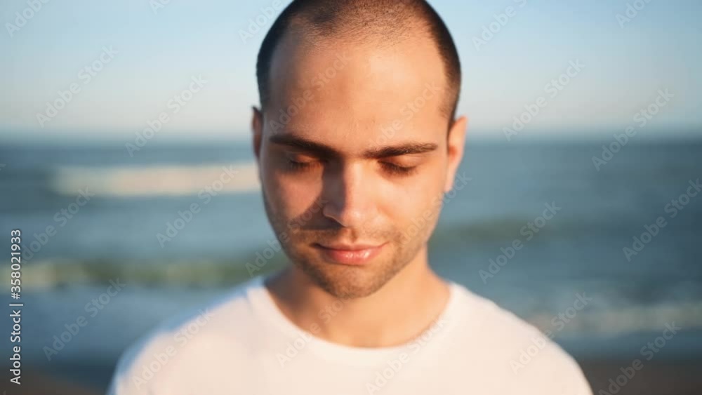 Portrait of young handsome man looking and smiling at camera standing on the beach with a sea on background. Happy pacified guy looking at camera relaxing on ocean shore on sunny day outdoors. Portrait of young handsome man looking and smiling at camera standing on the beach with a sea on background. Happy pacified guy looking at camera relaxing on ocean shore on sunny day outdoors.