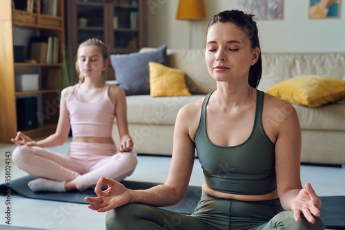 Calm middle-aged mother meditating with closed eyes together with daughter at home
