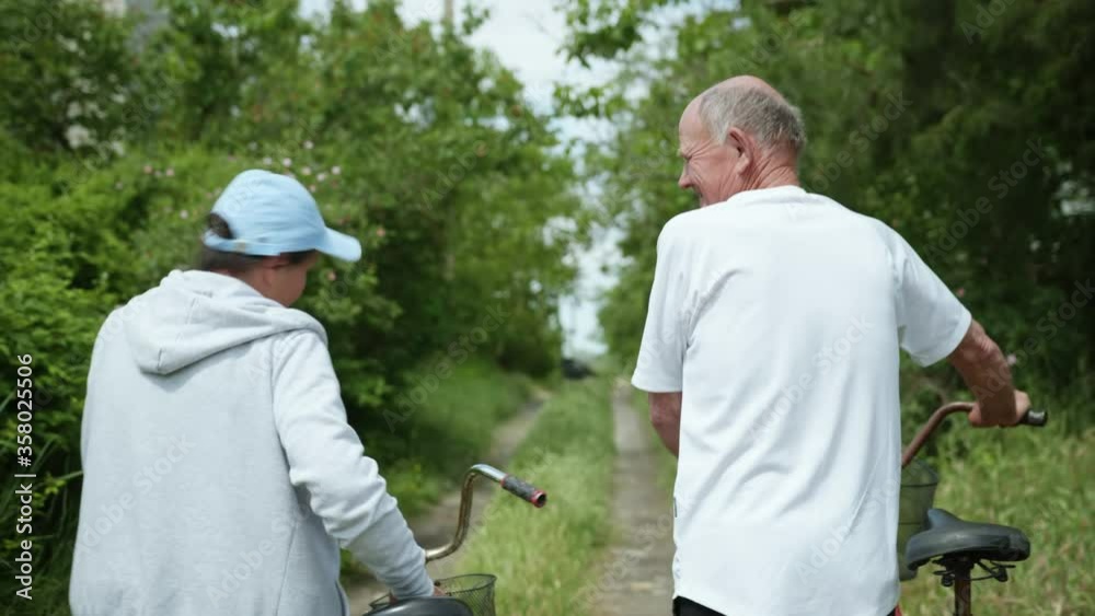 cheerful pensioners, an old man and an elderly woman have fun on walk with bicycles, cute grandparents happily chat on bike ride in countryside, rear view
