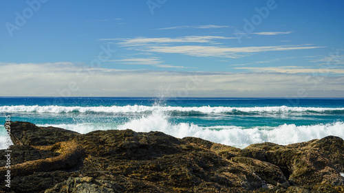 Looking out to sea with blue-green waves breaking on rocks in the foreground, and clouds on the horizon, beneath a blue sky. Currumbin, Gold Coast, Queensland, Australia