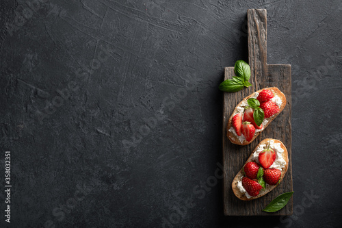bruschettas with strawberries, cream cheese and basil on a wooden board on a black background. Top view, place for text