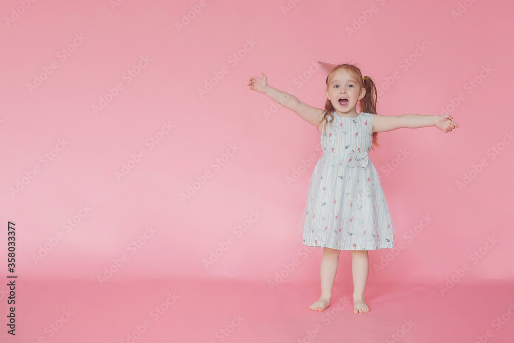Little cute happy girl in a birthday cap with a happy face rejoices raising his hands up on a pink background. Copy space