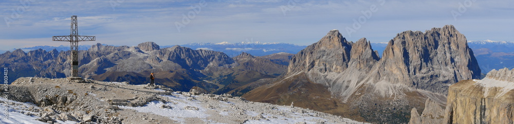 Panoramafoto mit Gipfelkreuz des Sass Pordoi, Sas de Pordoi in den Dolomiten, Südtirol, Italien, mit Blick auf die gegenüberliegende Bergkette sowie Langkofel und einem beobachtendem jungen Mann