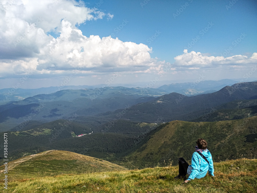 Girl sits on a mountainside and looks up at the sky. Natural summer landscape. Tourist traveling through the mountains. Green hills and mountains. Girl on a halt during a trip through the mountains.