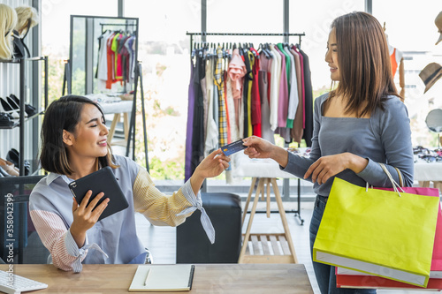 фотографія Young woman clothing shop owner holding tablet device receiving credit card payment from a female customer holding shopping bags inside a fashion clothing store