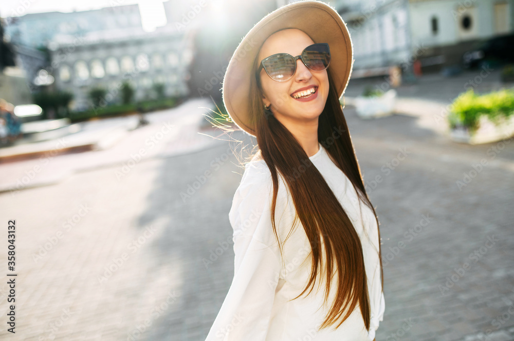 Cheerful beautiful brunette in a stylish hat and sunglasses looks at the camera and laughs happily. Stylish young woman with long hair walks around the city