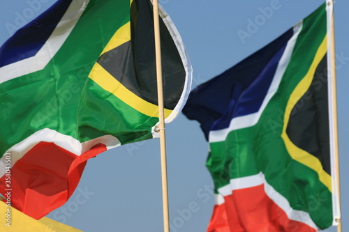 A couple of colourful South African flags blow in the wind whilst mounted onto thin stick poles set against a vivid cloudless blue sky taken during the summer season. London, England