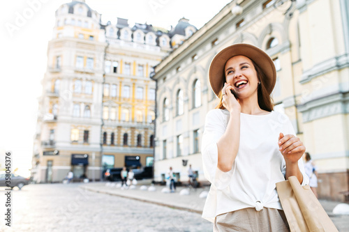 An attractive young girl is walking along a city street, talking on the phone and laughing. Close portrait of a stylish happy young woman in a hat and with a paper shopping bag