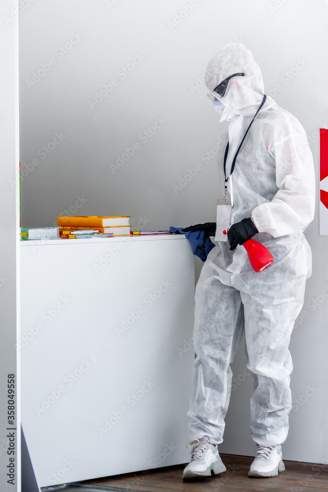 One medical worker in white protective suit wipes surface to disinfect cleaning public store place with professional detergents spray sanitizer to stop spreading virus infections Covid-19