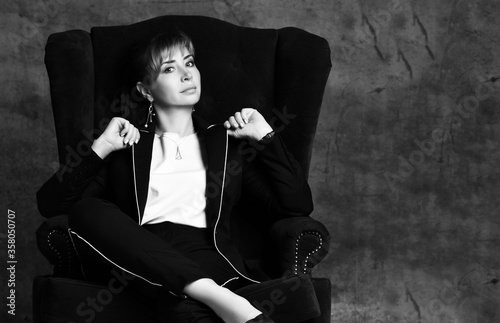 Black and white portrait of young beautiful woman in black formal business costume sitting in plush luxury armchair