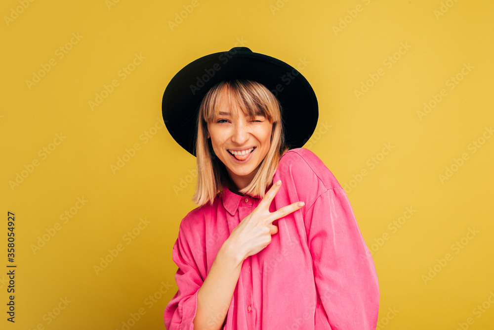 Cheerful happy positive young woman posing on camera with smile. Hold two fingers up in peace sign and keep one eye closed. Stylish trendy girl isolated over yellow background. Cheerful happy positive young woman posing on camera with smile. Hold two fingers up in peace sign and keep one eye closed. Stylish trendy girl isolated over yellow background.
