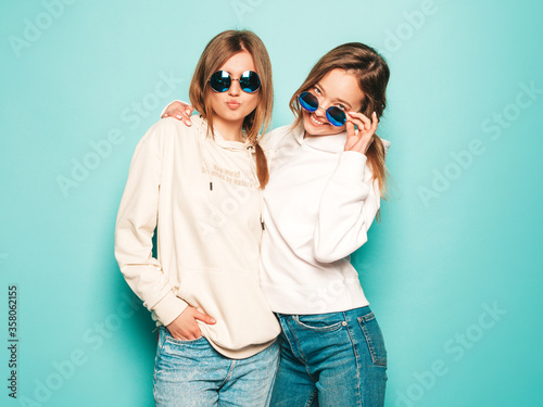 Two young beautiful blond smiling hipster girls in trendy summer hoodie clothes. Sexy carefree women posing near blue wall. Trendy and positive models having fun in sunglasses © halayalex